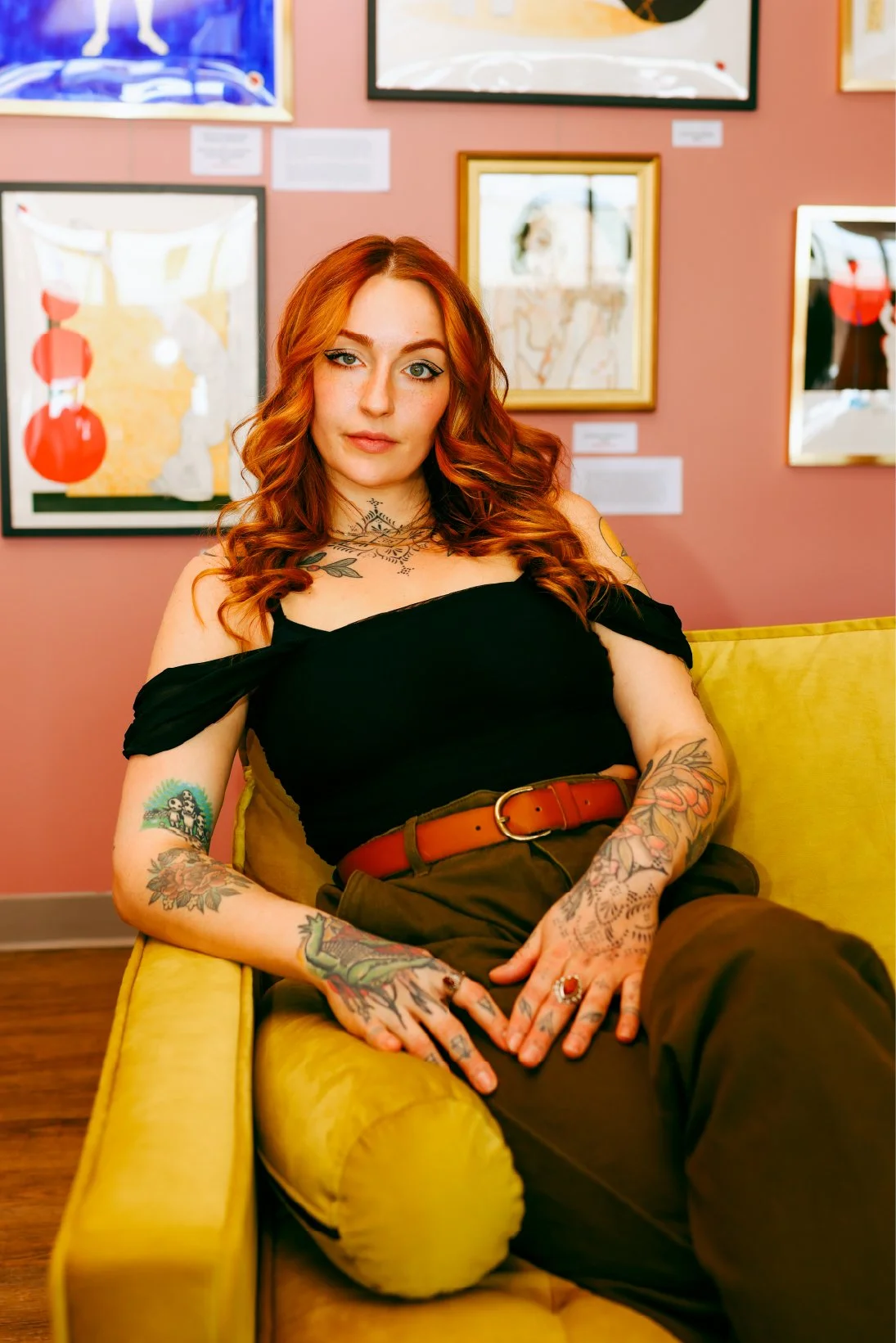 A woman with red hair and tattoos sitting on a yellow sofa in an art gallery, with colorful framed artwork on the pink wall behind her.