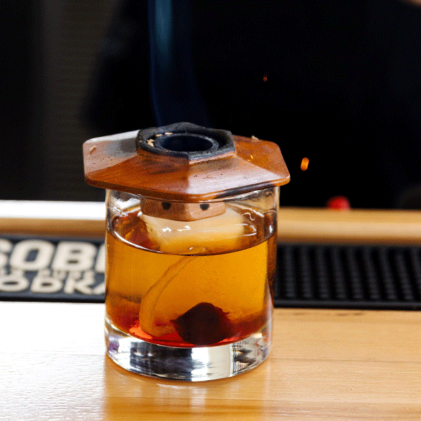 Cocktail with a lemon slice and a cherry in a glass, with a burning wooden piece on top