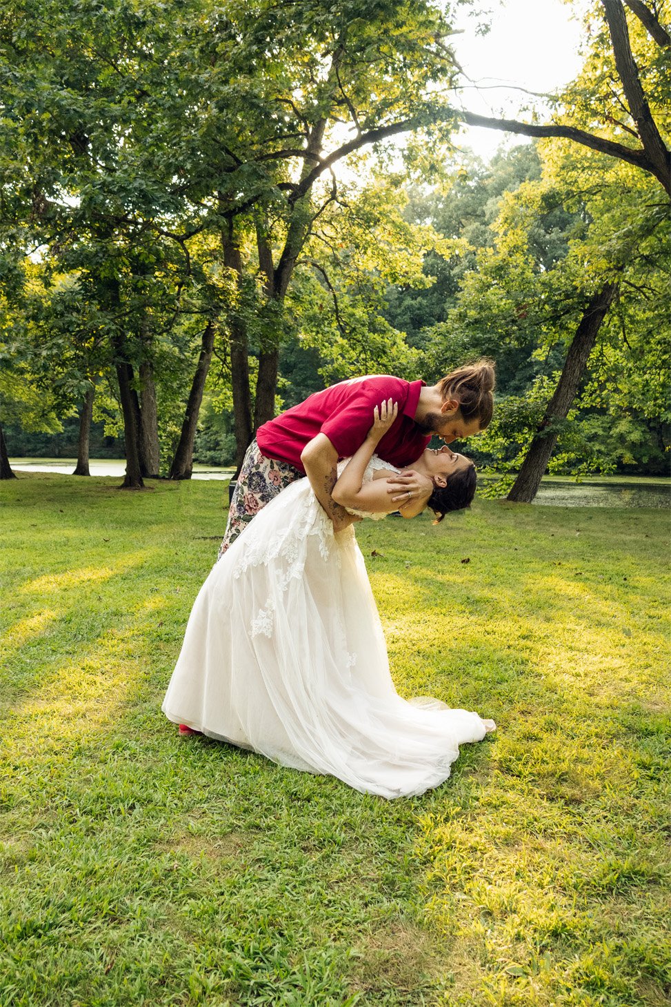 A couple dressed in wedding attire sharing a romantic moment outdoors in a park with green trees and grass.