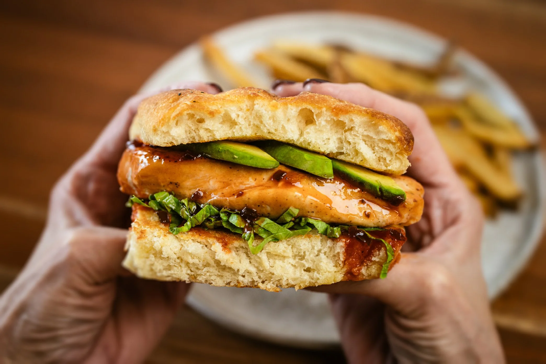 A close-up of a hand holding a sandwich with grilled chicken, green avocado slices, chopped green herbs, and a dark sauce on a toasted bun, with a side of French fries in the background.