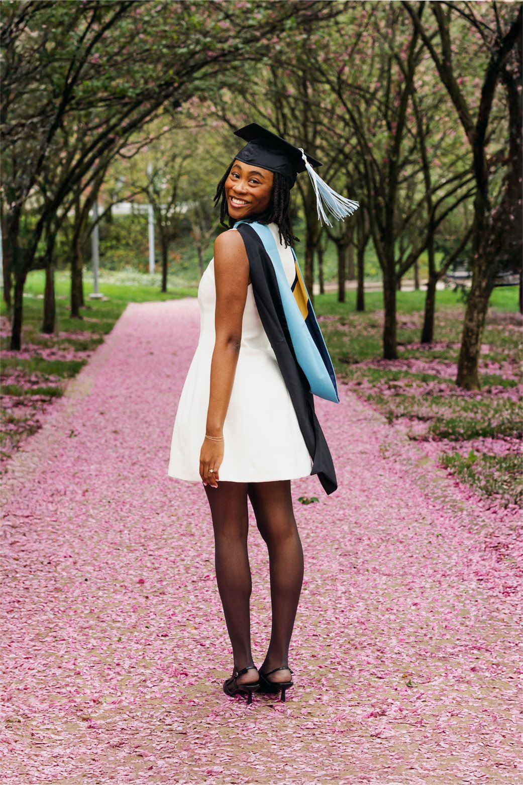 A smiling woman in a white dress, wearing a graduation cap and gown with honor cords, stands on a pink petal-lined pathway in a park with trees and pink flowers overhead.