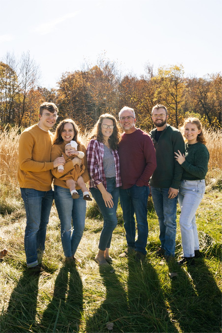 Family picture outdoors on sunny autumn day with seven people, including a baby, standing on grass with tall trees showing fall foliage in the background.
