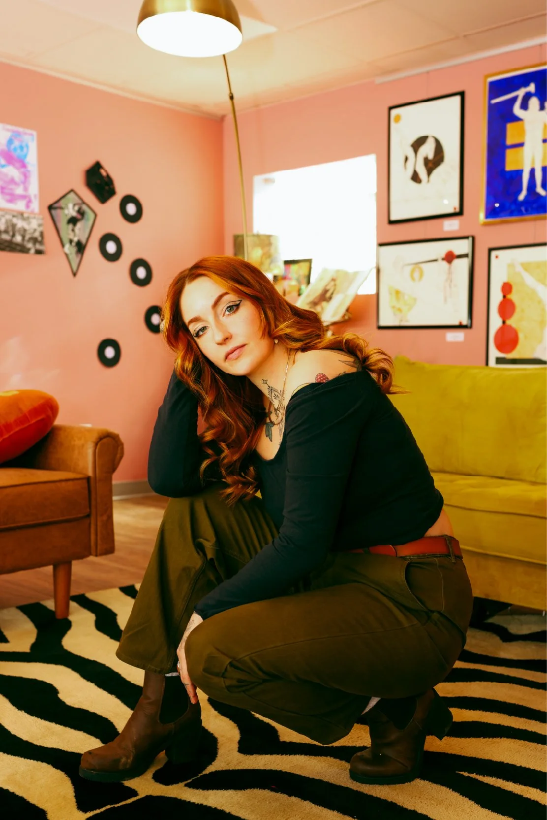 A woman with long red hair and tattoos, dressed in a black top and olive green pants, crouches on a zebra-patterned rug in a colorful art-filled room.