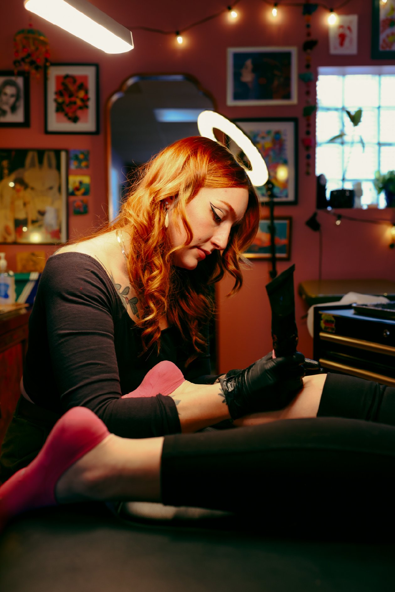 A woman with red hair and a black outfit, wearing black gloves and red shoes, sitting on a table in a colorful, art-filled room. She appears to be tattooing or drawing on someone's leg, which is partially visible in the foreground. The room has pink 