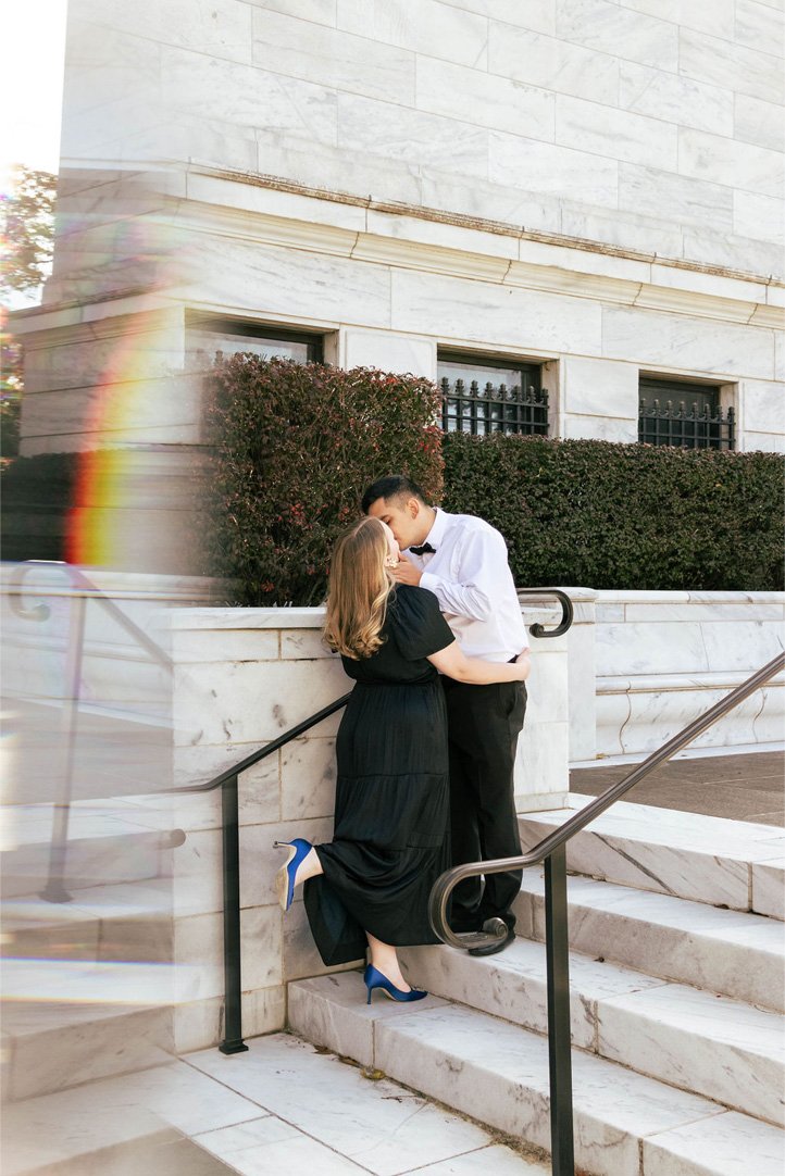 A couple dressed formally, with the woman in a black dress and blue heels and the man in a white shirt and black pants, sharing a kiss on the steps outside a marble building.