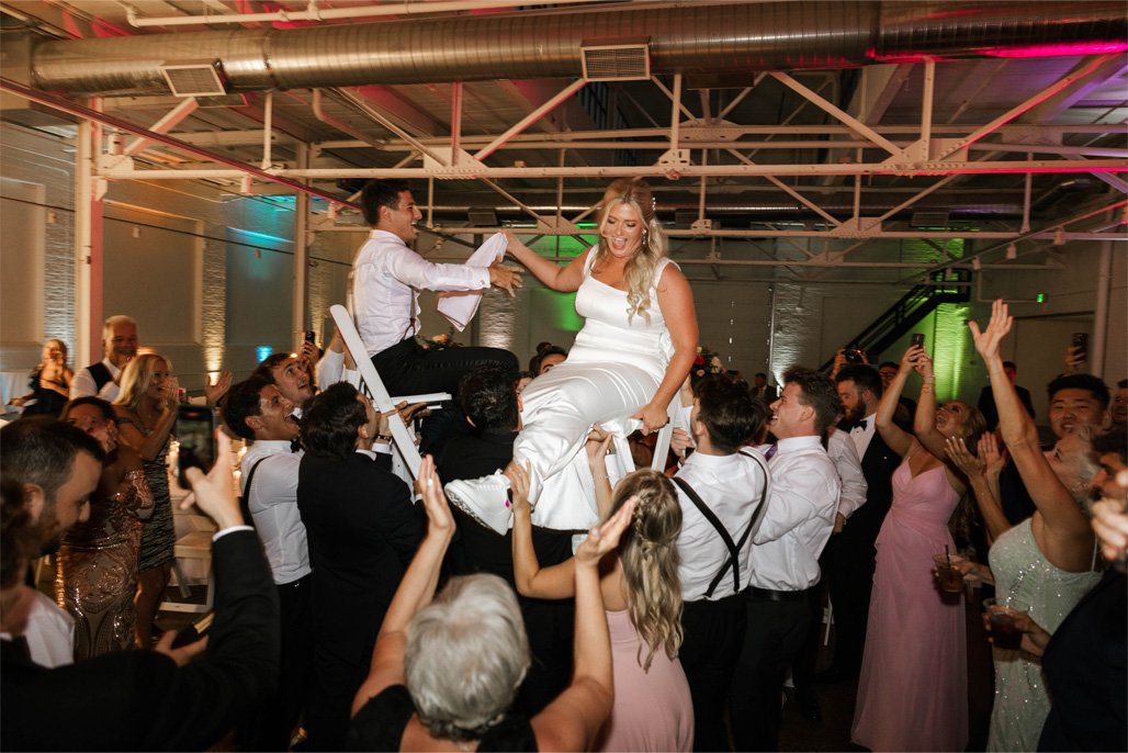 A bride in a white dress is being lifted on a chair by a group of guests at a celebration or wedding reception. Guests are dancing and clapping around her in a decorated indoor venue.