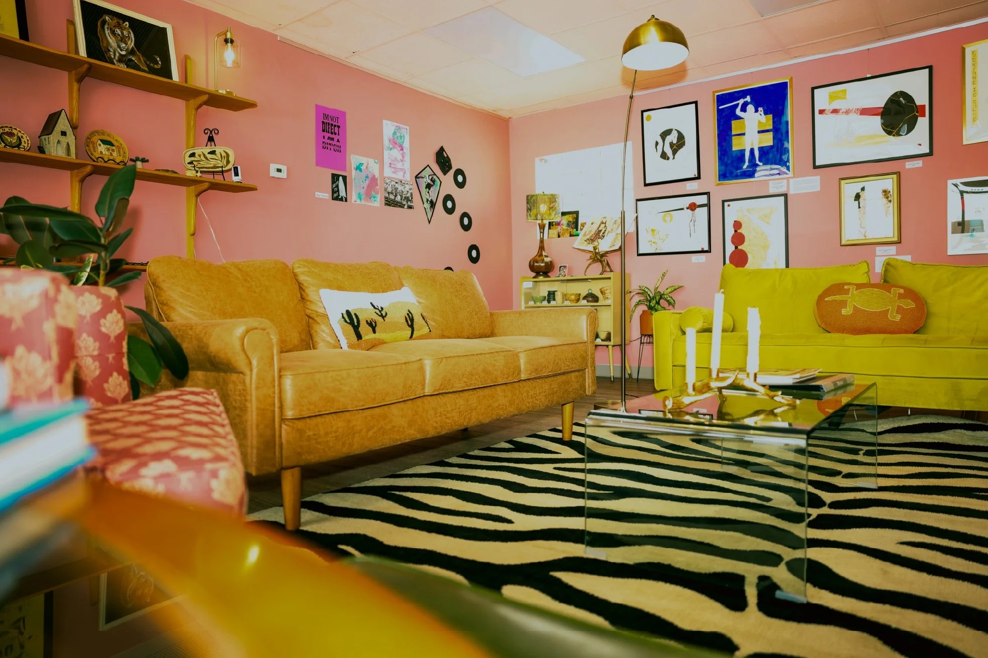 Cozy living room with pink walls decorated with framed art, a mustard yellow couch, a yellow armchair, plants, a zebra-patterned rug, and various decorative items and artwork on the walls.