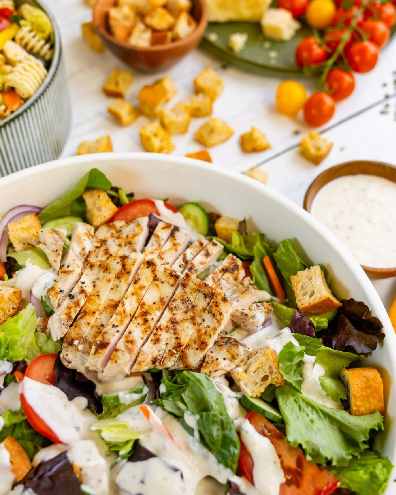 Close-up of a fresh grilled chicken salad with lettuce, cherry tomatoes, cucumbers, croutons, and creamy dressing in a white bowl. In the background, there are tomatoes, croutons, and dipping sauce on a white table.