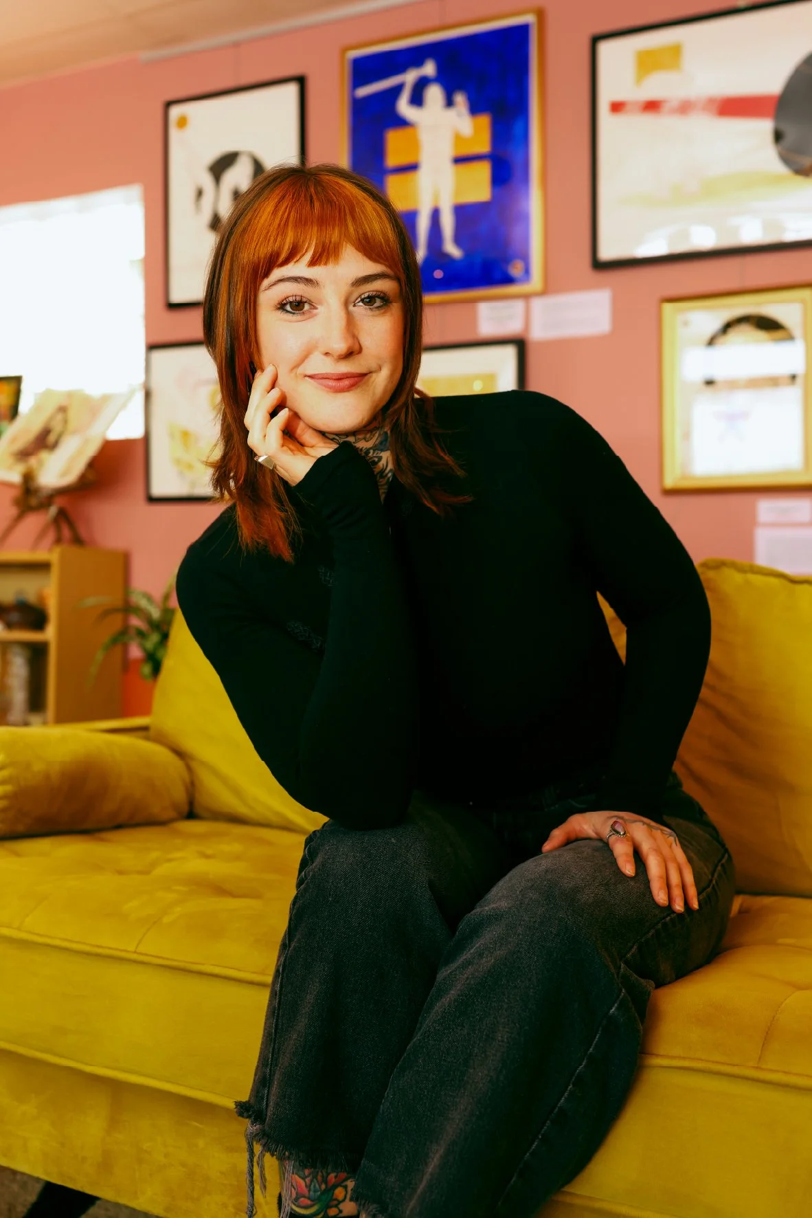 A young woman with red hair sitting on a yellow couch, smiling, in a colorful art gallery with framed artworks on the pink wall behind her.