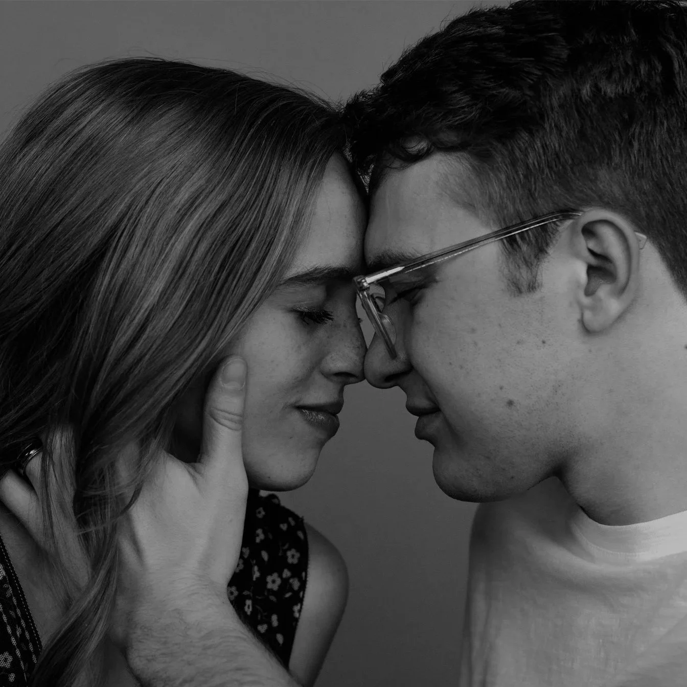 Close-up black-and-white photo of a young couple touching foreheads with eyes closed; woman with long hair and a man wearing glasses, both conveying emotion.