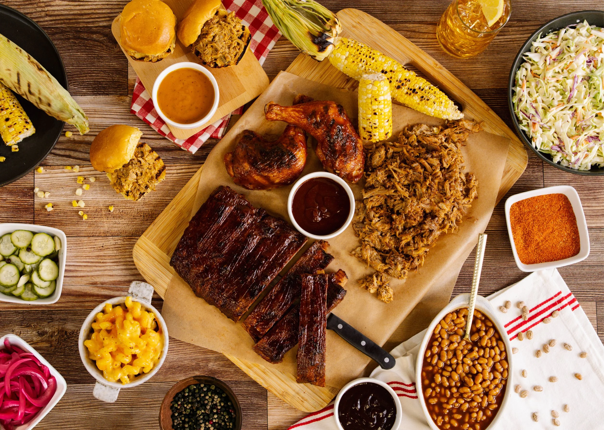 A spread of barbecue food on a wooden table, including corn on the cob, pulled pork, ribs, slider sandwiches, coleslaw, baked beans, macaroni and cheese, and various sauces and side dishes.