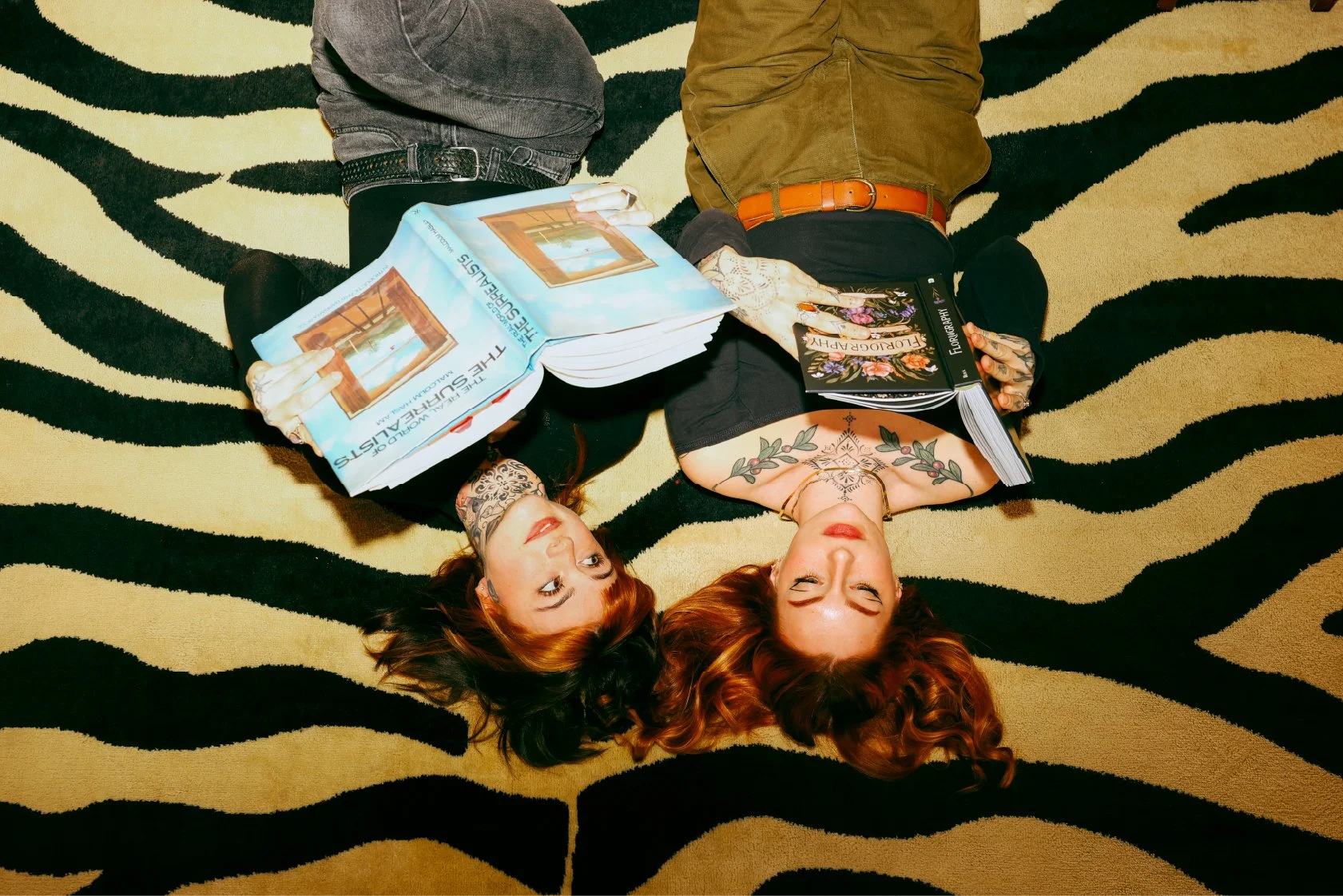 Two women with red hair and tattoos lying on a black and beige zebra print rug, each holding a book and looking up at the camera. The woman on the left has a black top and black jeans, holding a self-help book titled 'The Subtle Art of Not Giving a F