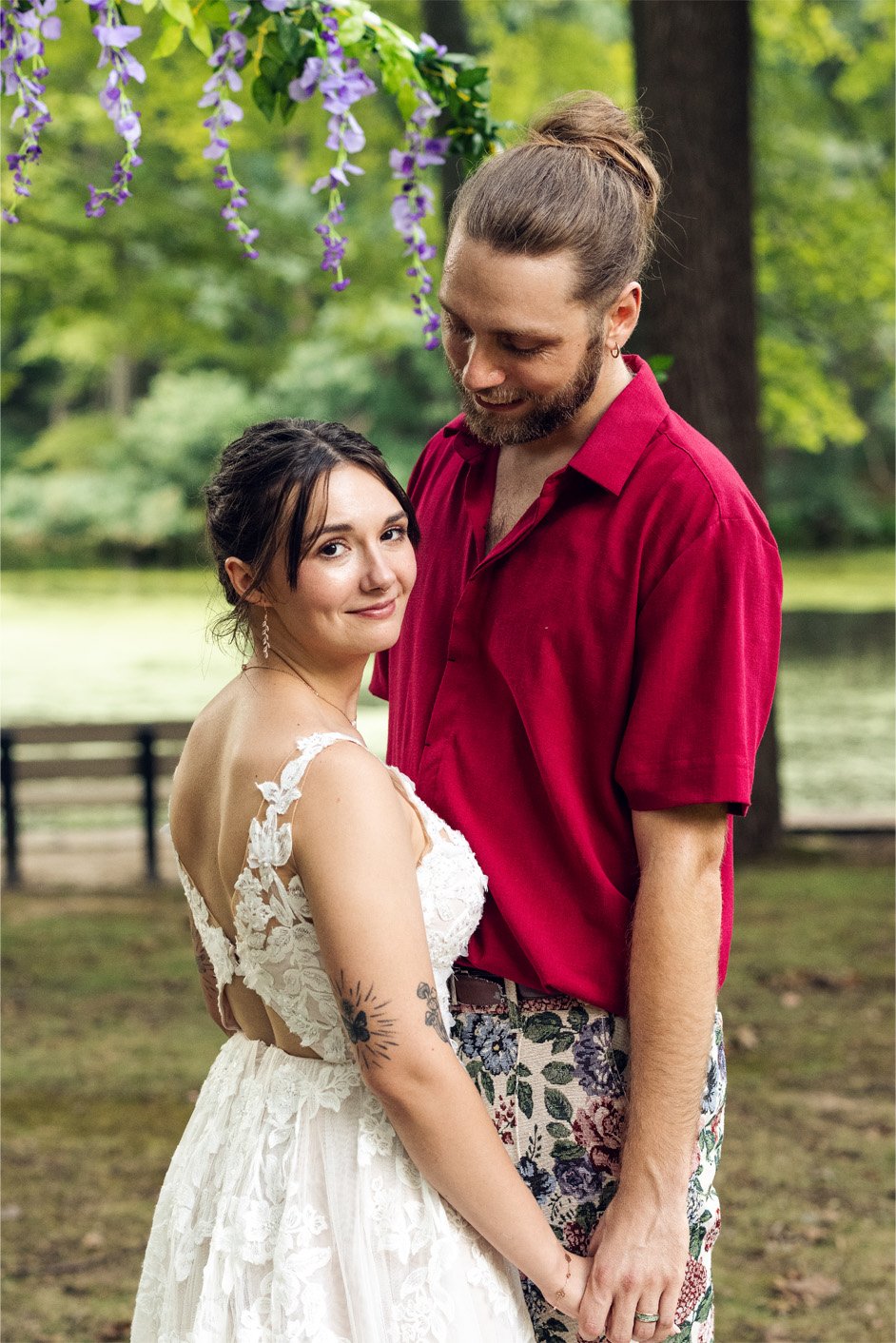 A couple standing outdoors near a lake, holding hands and smiling. The woman is in a white lace dress with tattoos on her arm, and the man is in a red shirt with floral patterned pants. There are trees and purple wisteria flowers overhead.