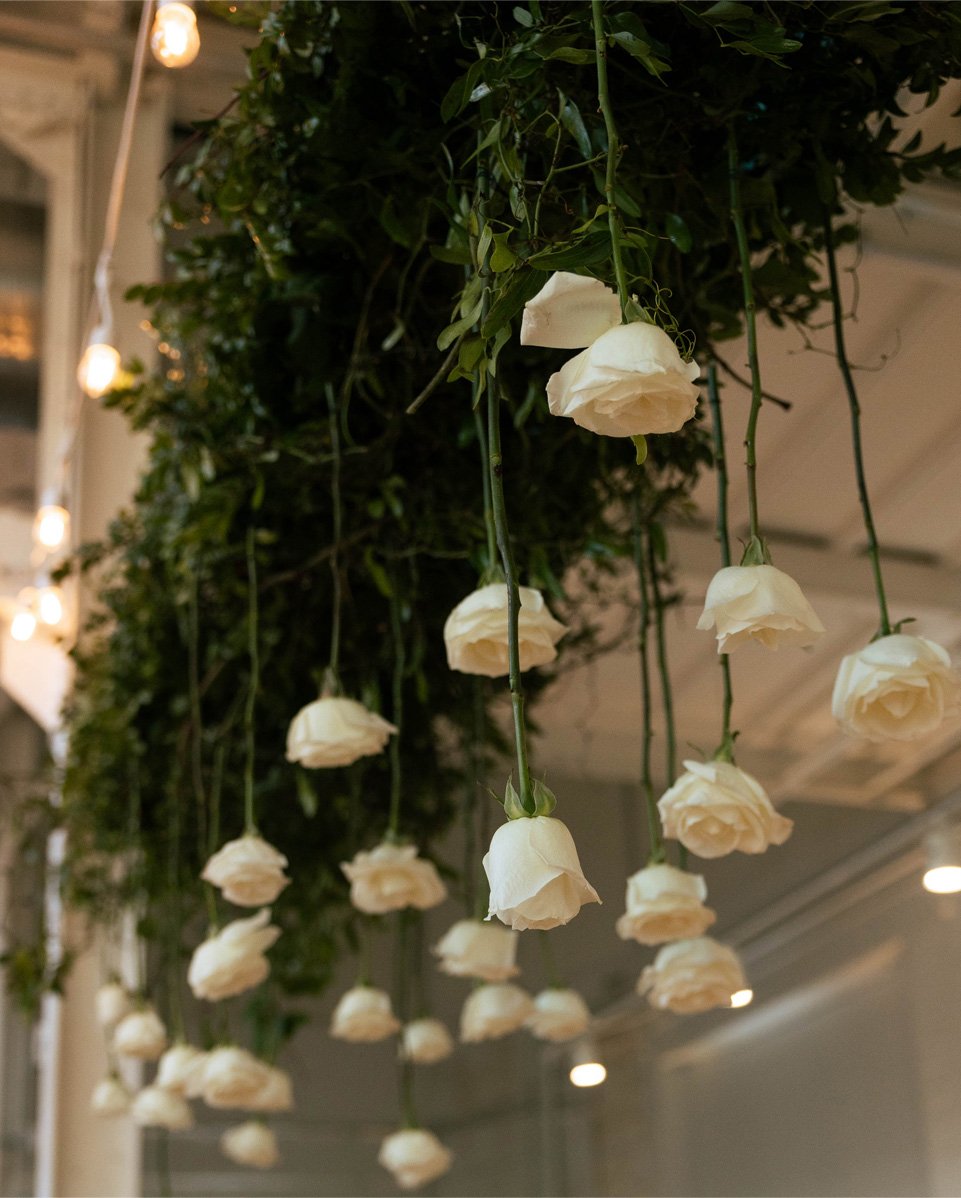 Hanging white roses and greenery decoration inside a building with string lights and ceiling panels.