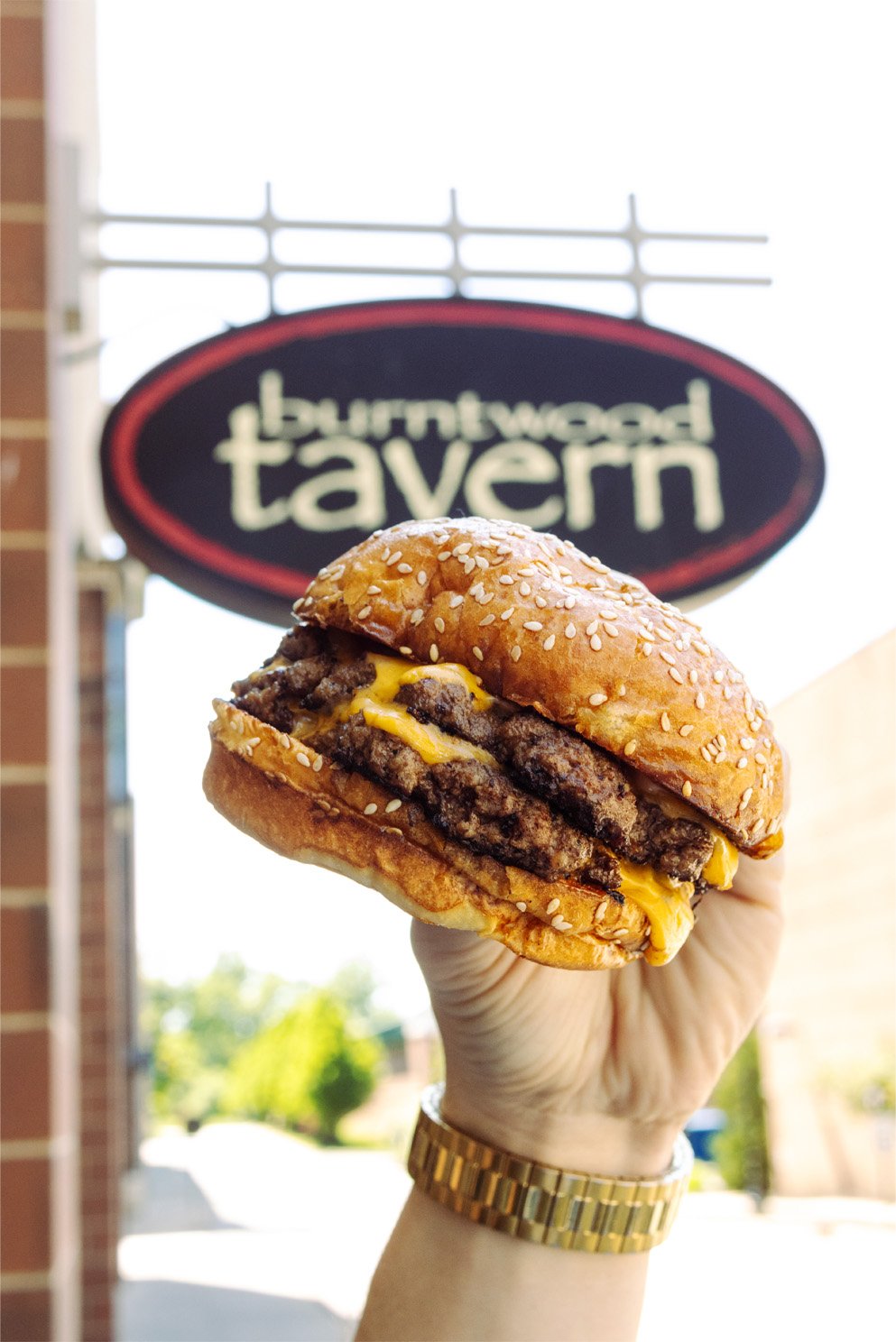 A hand holding a cheeseburger with multiple beef patties, melted cheese, and a sesame seed bun in front of a sign that reads 'Buntwood Tavern'.