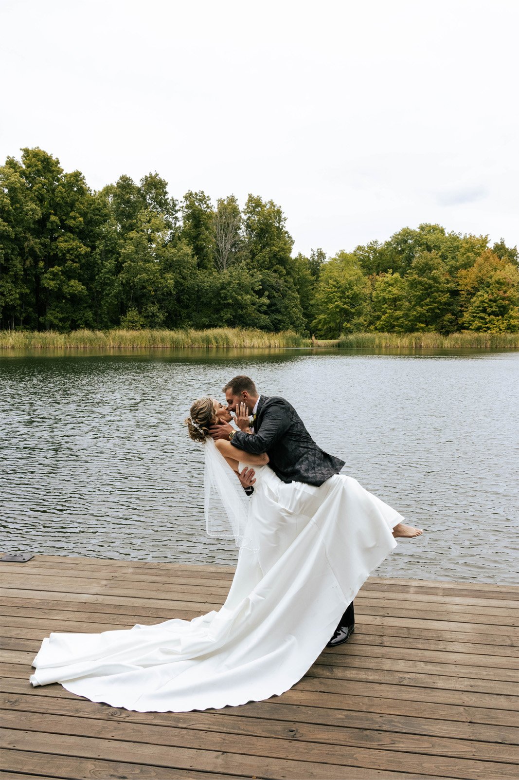 A groom in a black suit dips a bride in a white wedding dress for a kiss on a wooden dock by a lake, surrounded by green trees under an overcast sky.