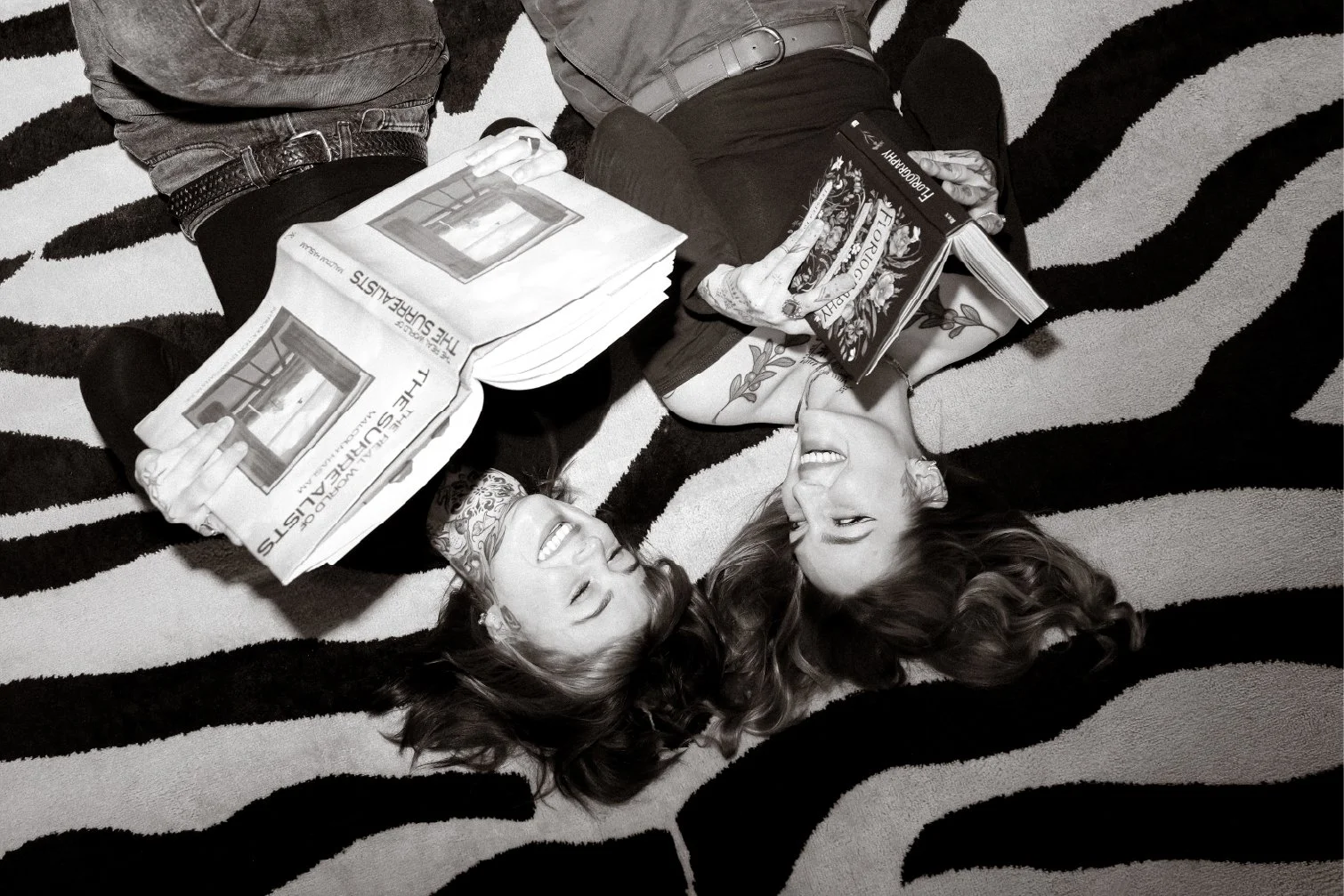 Two women lying on a patterned rug, smiling and holding books, engaging in conversation.