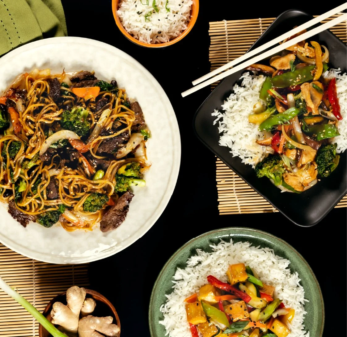 A variety of Asian dishes including stir-fried noodles with vegetables and beef, vegetable stir-fry over rice, and mixed vegetables served with rice, with bowls and chopsticks on a black table.