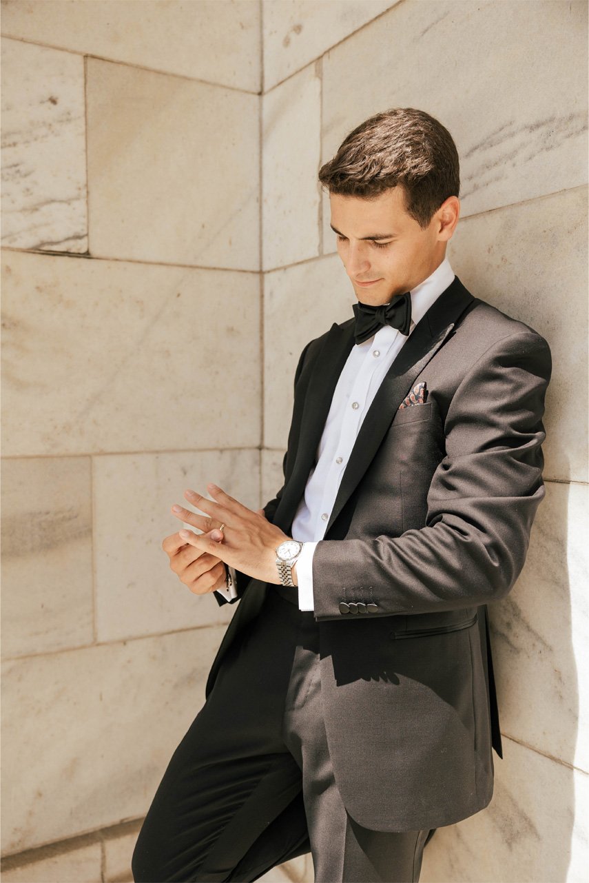 A man in a black tuxedo with a bow tie, wearing a watch, leaning against a stone wall and looking at his hands.