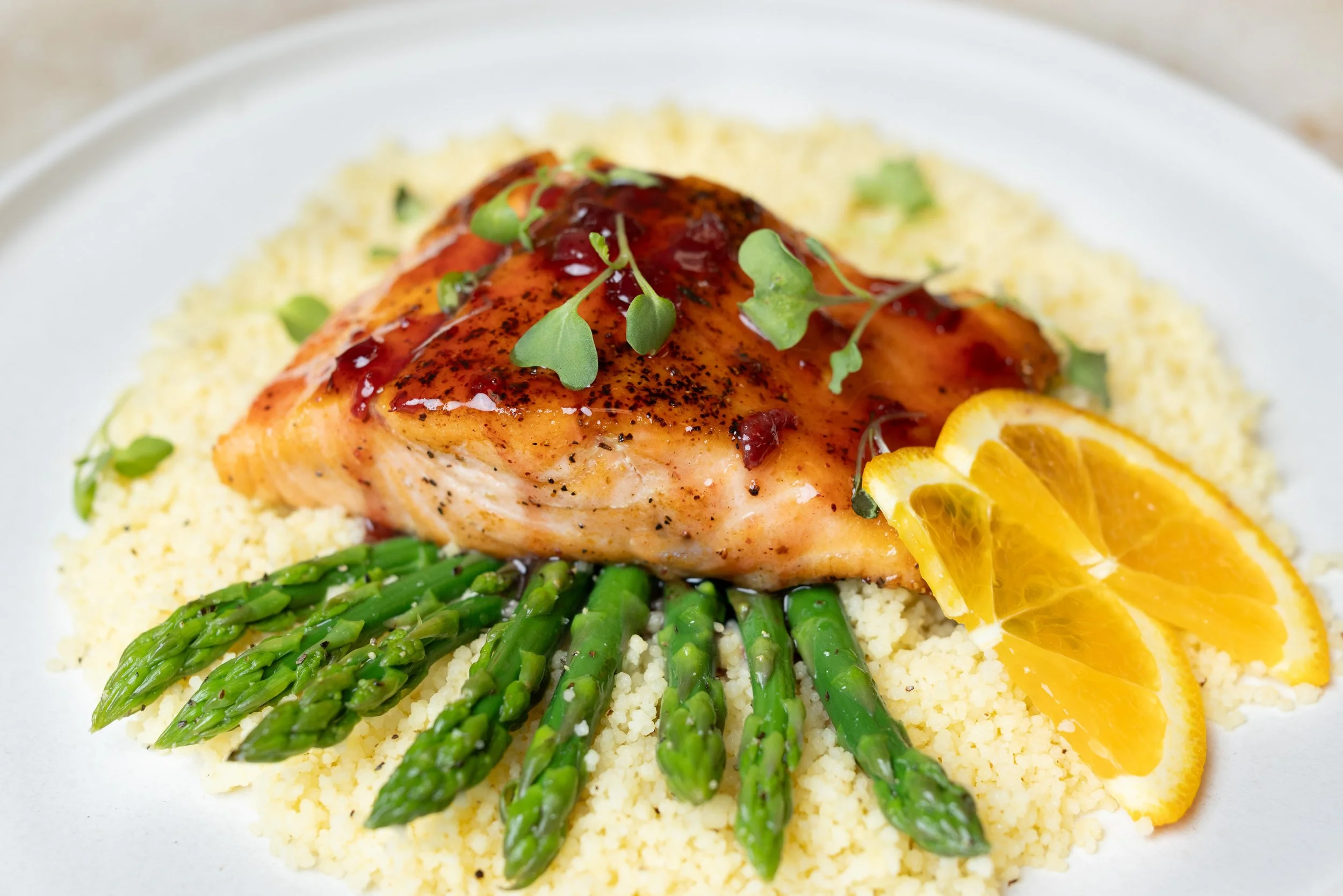 A plate of cooked salmon topped with sauce and microgreens, served with lemon slices, asparagus, and a side of mashed potatoes or couscous.