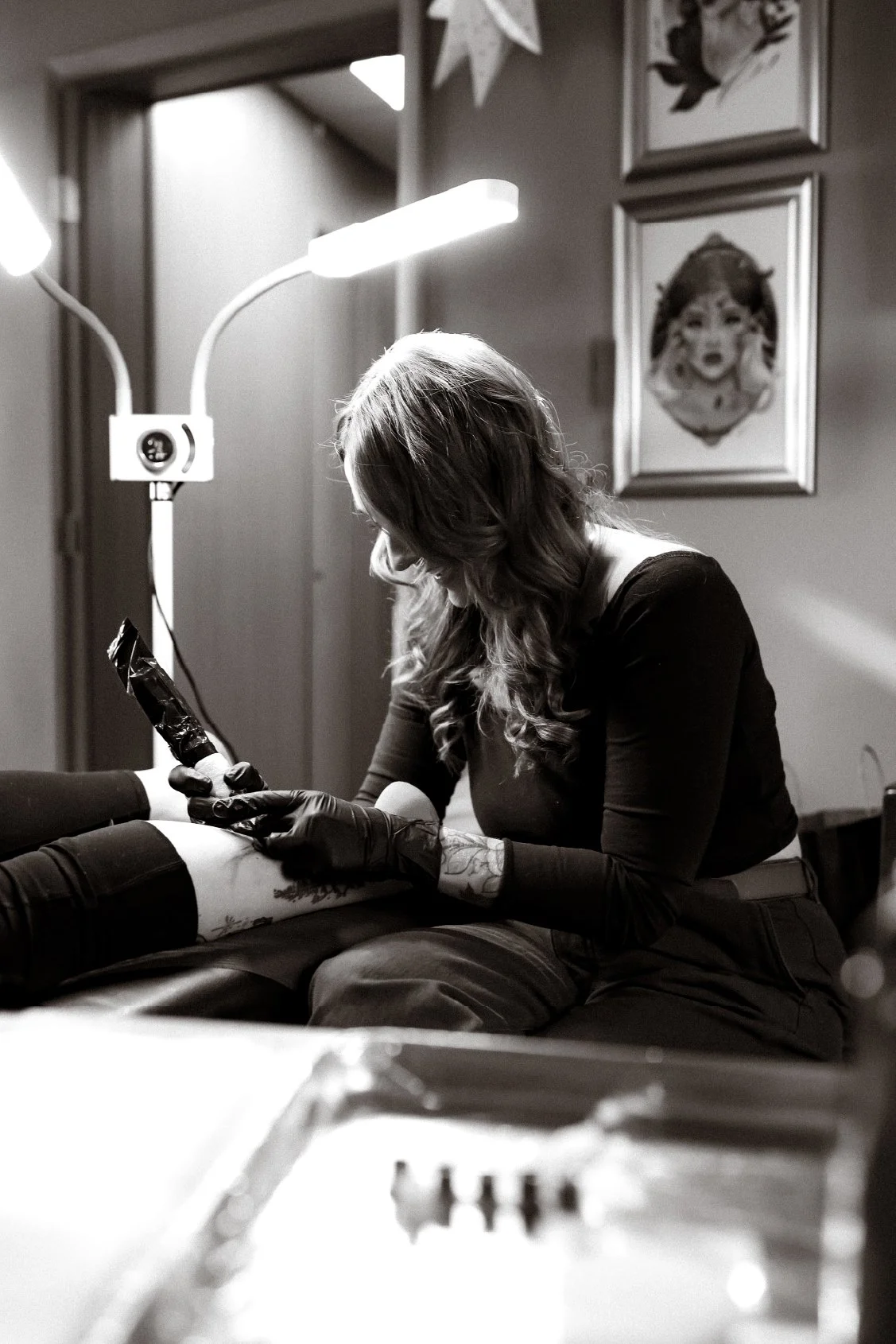 A woman with wavy hair wearing a dark shirt and gloves, sitting on a tattoo chair, getting a tattoo on her leg in a tattoo studio.