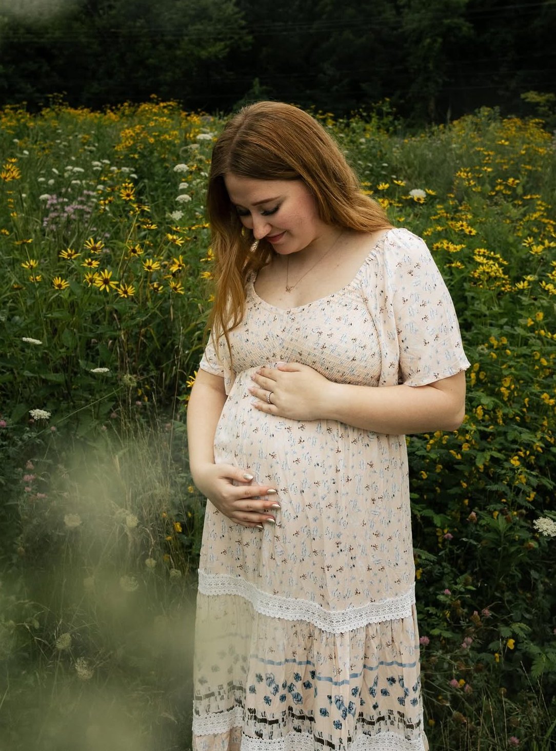 Pregnant woman in a floral dress standing in a field of yellow flowers, gently holding her baby bump.