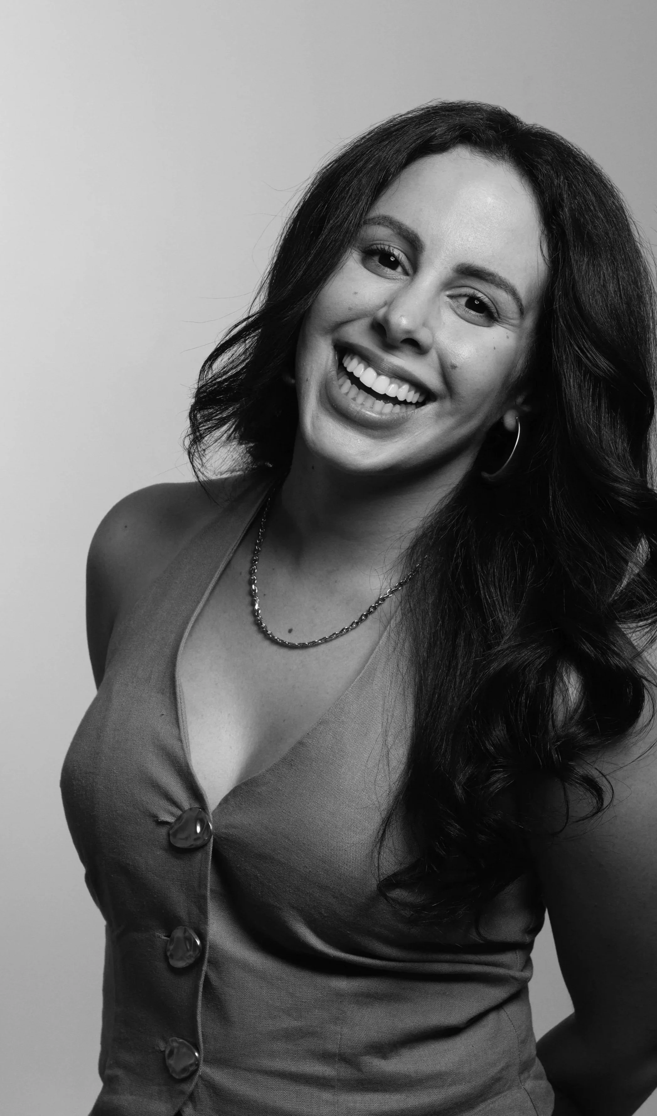 A black-and-white photo of a smiling woman with long wavy hair, wearing a sleeveless dress, hoop earrings, and a chain necklace, standing against a plain background.