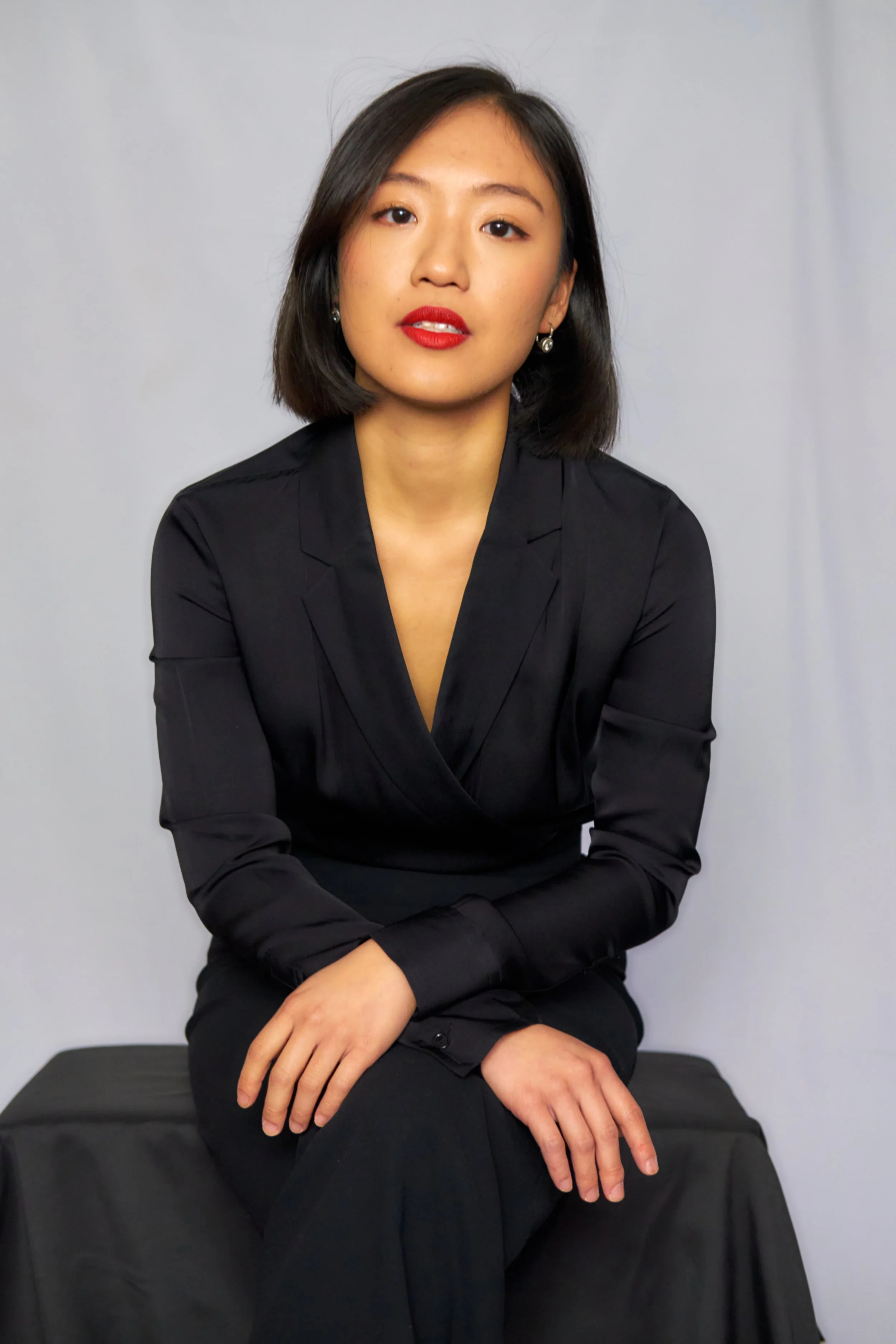 A woman with shoulder-length black hair, red lipstick, and earrings, sitting with crossed hands on her lap against a light grey background, wearing a black blazer.