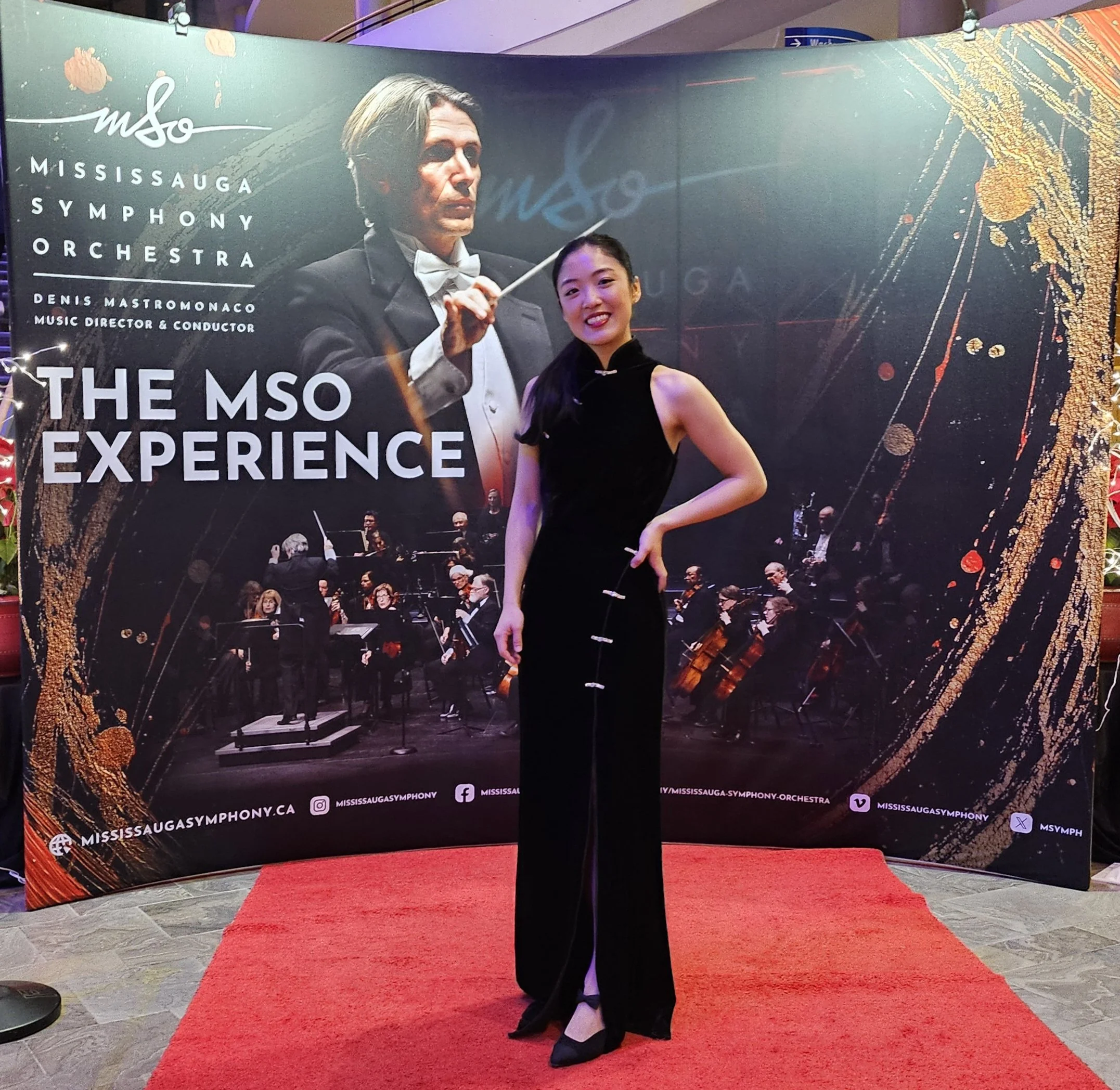 A woman in a black dress kneeling on a red carpet in front of a large promotional backdrop for the Mississippi Symphony Orchestra. The backdrop features a conductor in tuxedo with a baton and arrangements of musical notes and gold decorative elements.