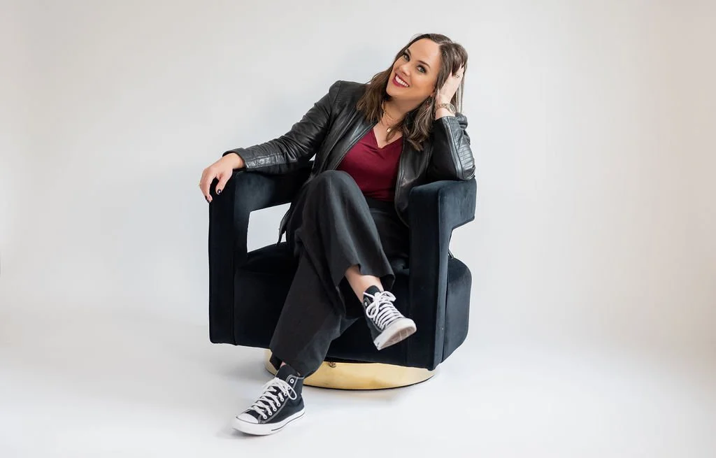 Cara Chace, a business coach for female entrepreneurs, sitting on a black chair in a black leather jacket and converse sneakers, smiling and inviting other women entrepreneurs to hire a business coach