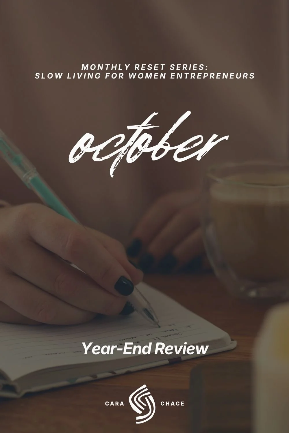 A hand with dark nail polish writes in a notebook beside a glass mug of coffee, with the words october and Year-End Review.