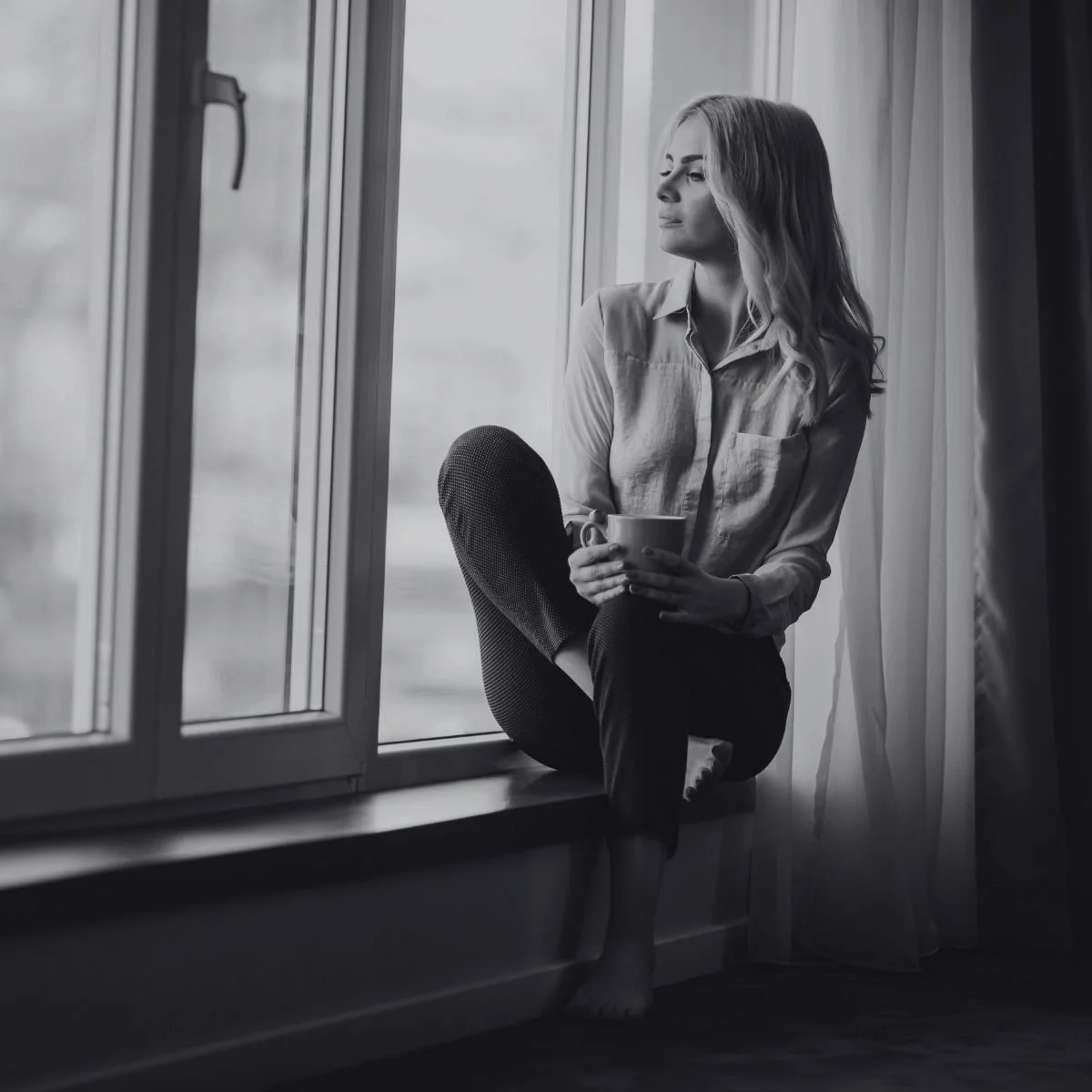 A woman sits on a window ledge holding a mug, looking outside in black and white, reflecting on her business year.
