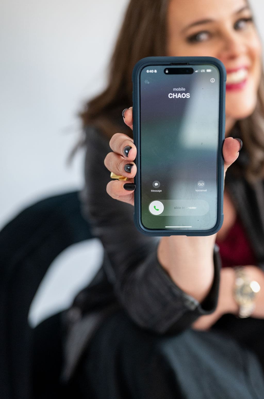 Cara Chace - The Productivity Rebellion free monthly guide - woman holding a smartphone with the screen displaying an incoming call from 'CHAOS'. She has dark nail polish and is smiling, wearing a black leather jacket.