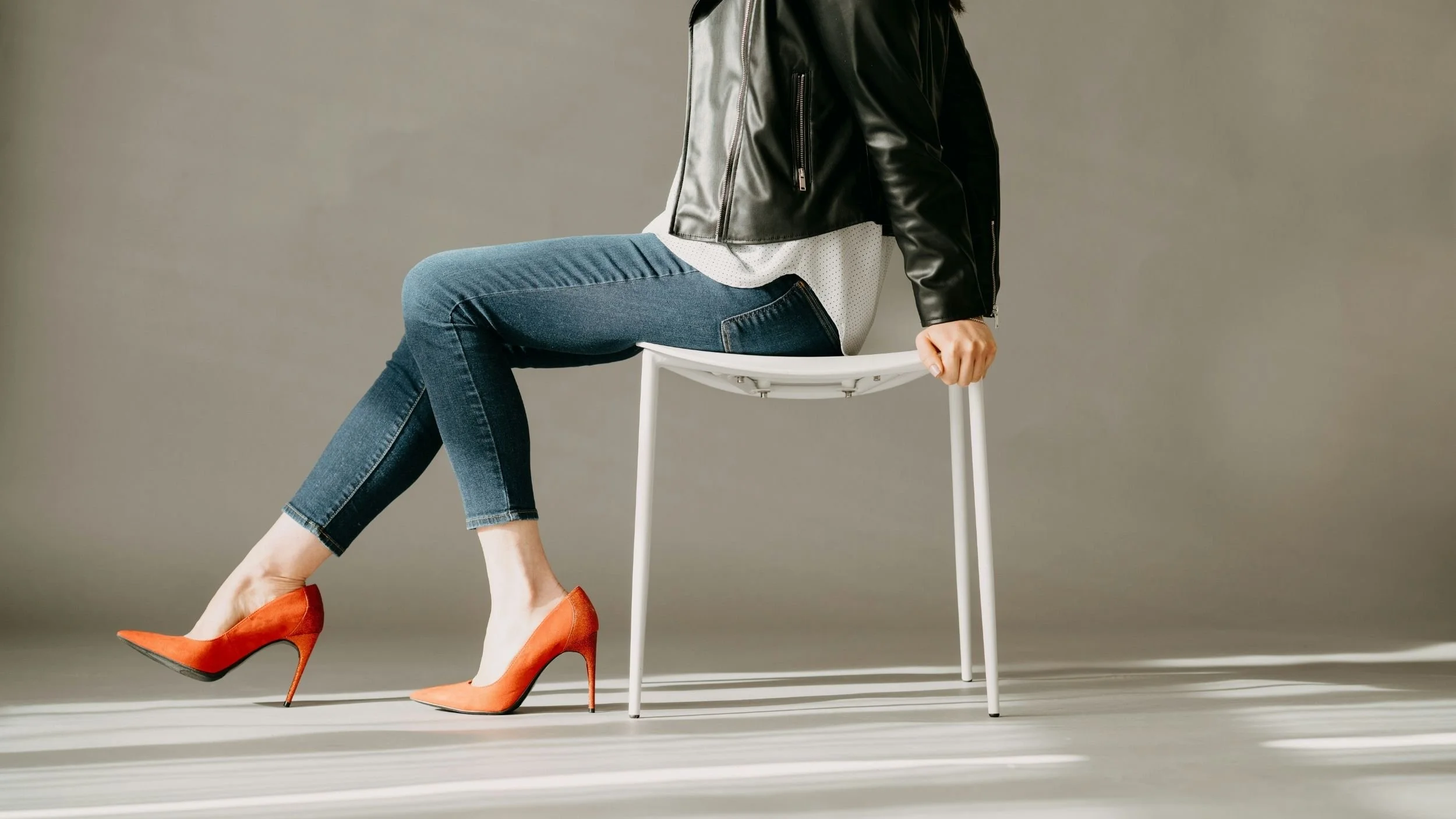 A woman wearing orange high heels, blue jeans, a white shirt, and a black leather jacket, sitting sideways on a white chair in front of a plain gray background - for The Productivity Rebellion free monthly guide