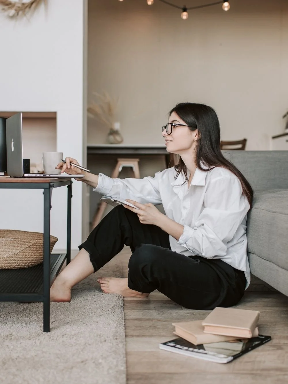 Woman entrepreneur sitting on the floor at home, notebook in hand, laptop open on a side table, looking up thoughtfully — coaching for overwhelmed women entrepreneurs.