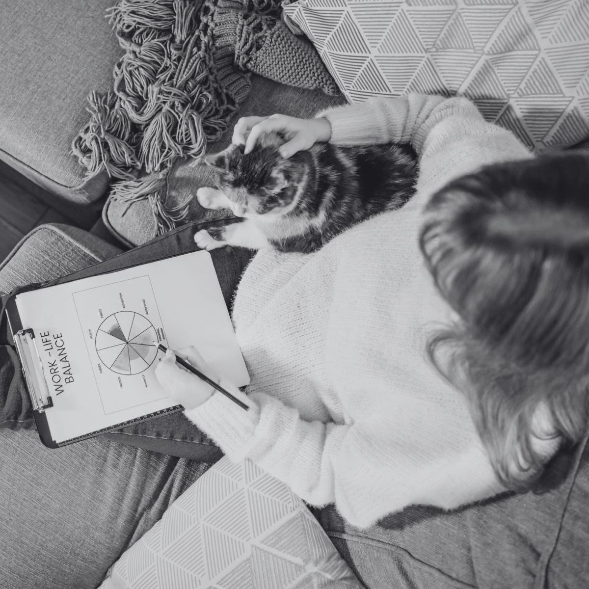 Woman on a couch petting a cat while filling out a work-life balance wheel worksheet on a clipboard