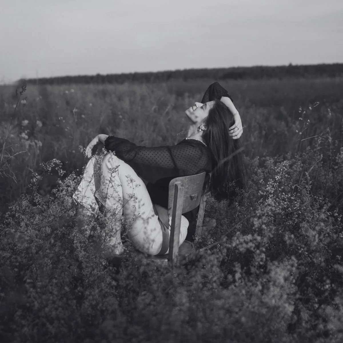 A woman rests back on a wooden chair in a wildflower field, eyes closed, in black and white — a slow reset.