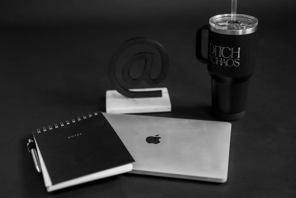 Dark flatlay with notebook, laptop, @ symbol decor, and Ditch the Chaos water tumbler, showing office supplies for a woman entrepreneur who needs business coaching