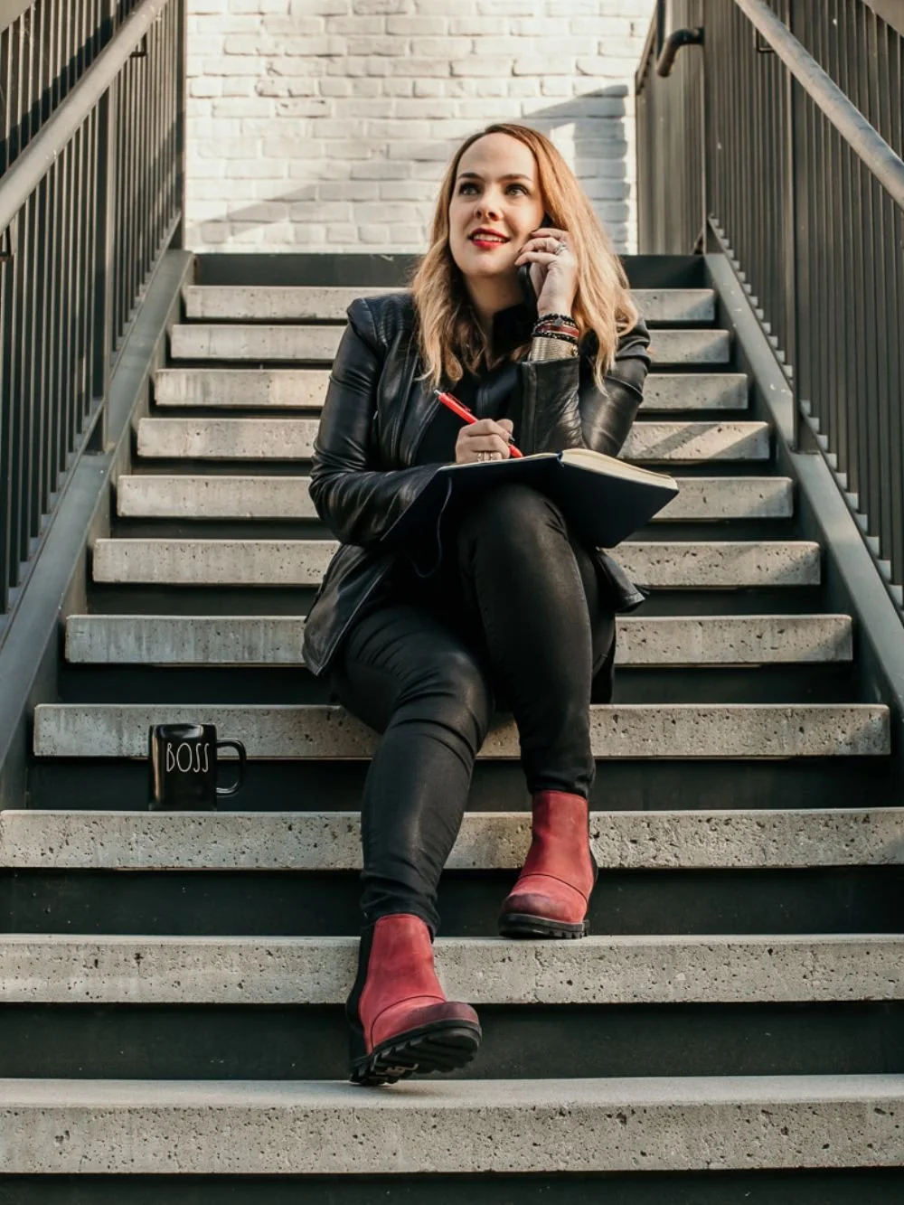 Cara Chace, a female business coach for entrepreneurs, sitting on steps on the phone with a notebook and "boss" coffee mug