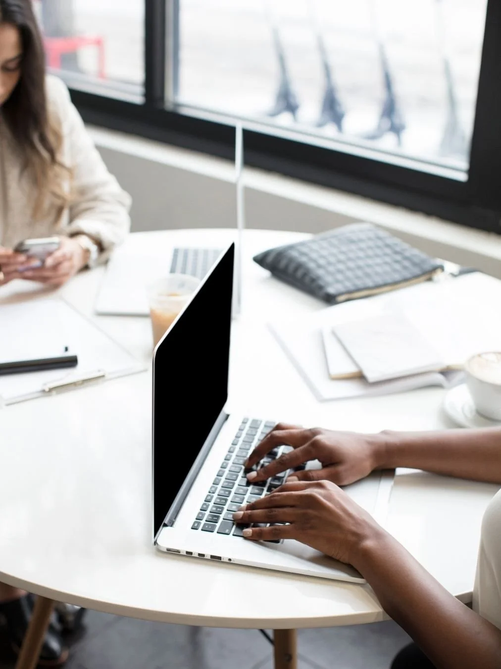 Two women entrepreneurs at a light and bright table working on phone and laptops, reflecting working with a business coach