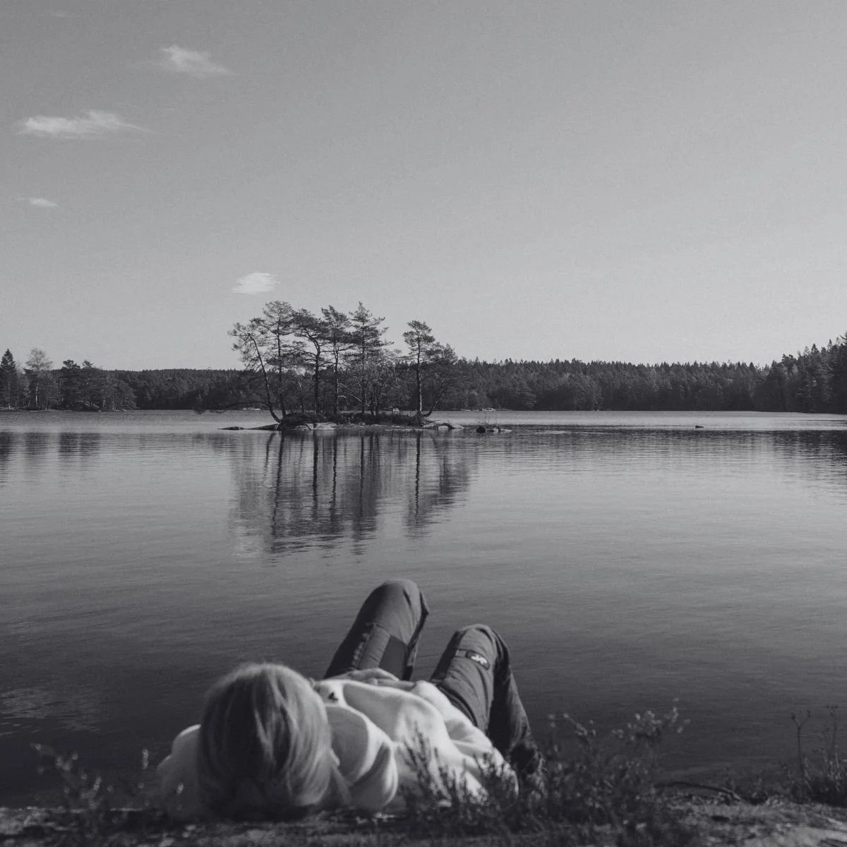A woman lies on a lakeshore in black and white, looking out at a calm lake and a small tree island.