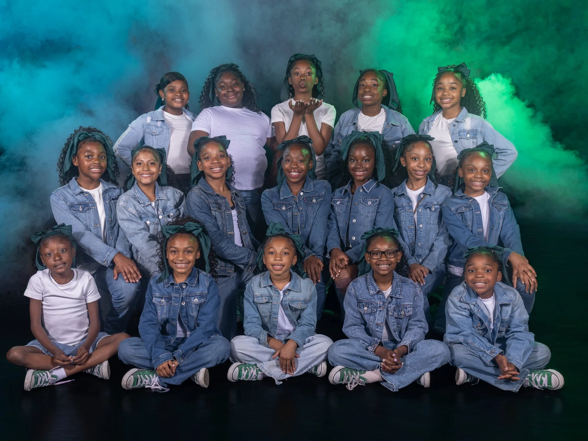 A group of young girls sitting and standing together in front of a green and blue smoky background, smiling and posing for a photo, wearing casual denim and white clothing.