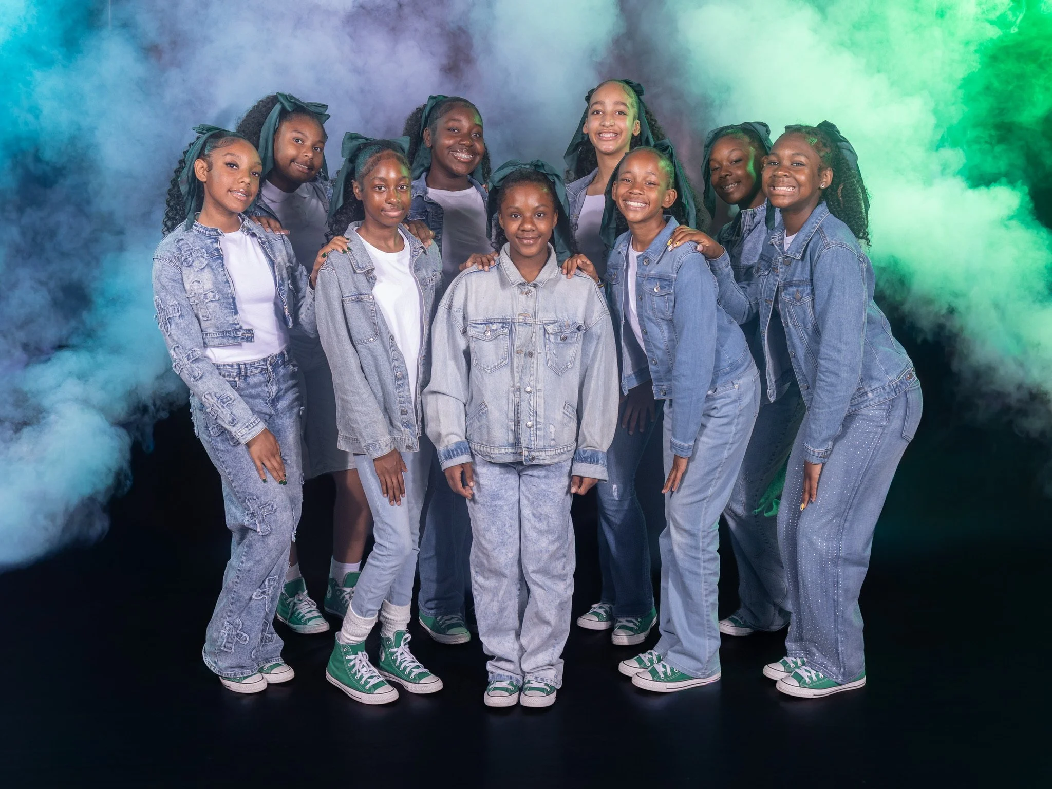 A group of ten young girls wearing denim jackets, white shirts, and green high-top sneakers, posing together against a colorful smoke background.