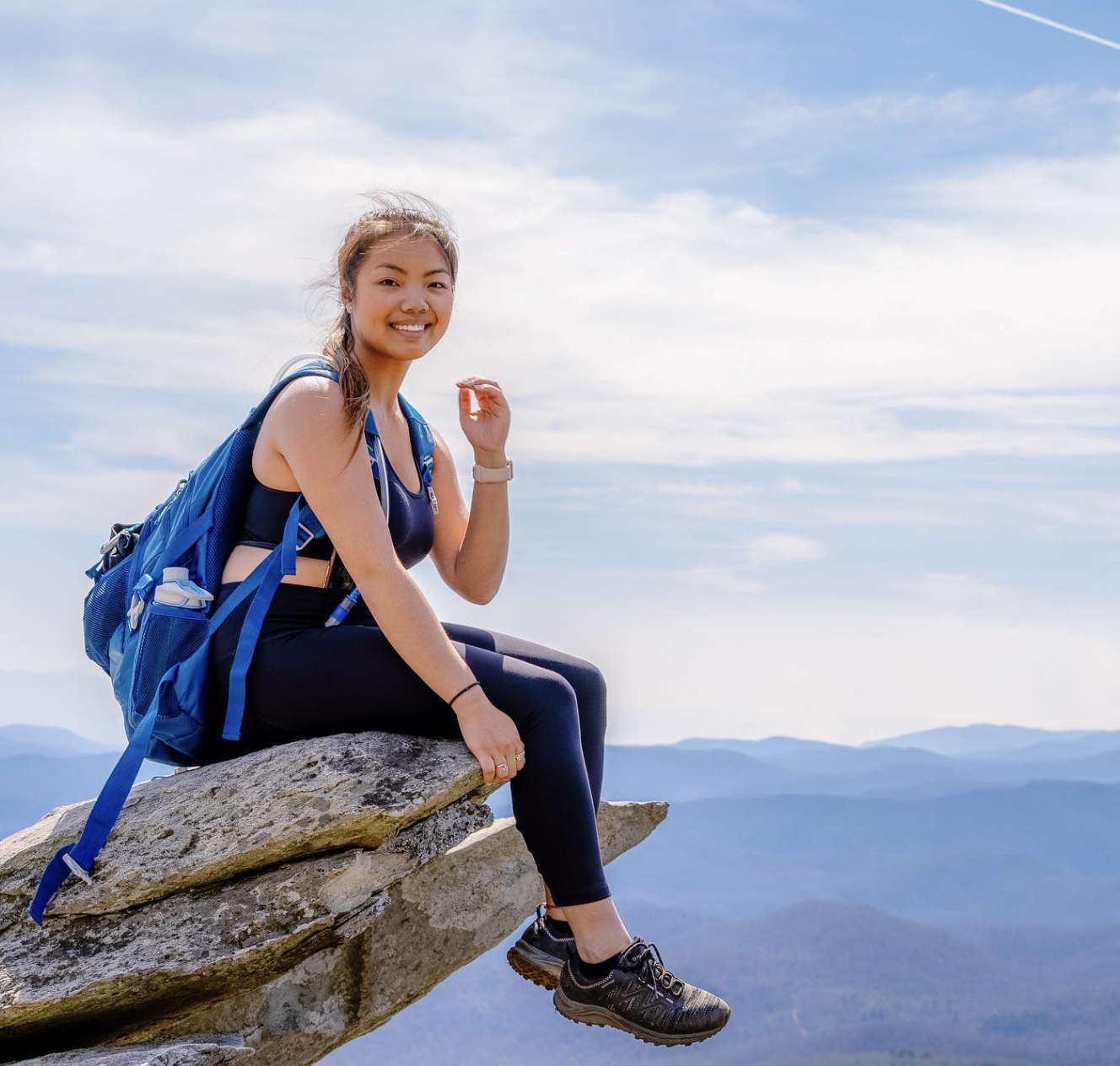 Woman sitting on edge of rock, wearing a blue backpack, looking at camera, with mountains and sky in background.