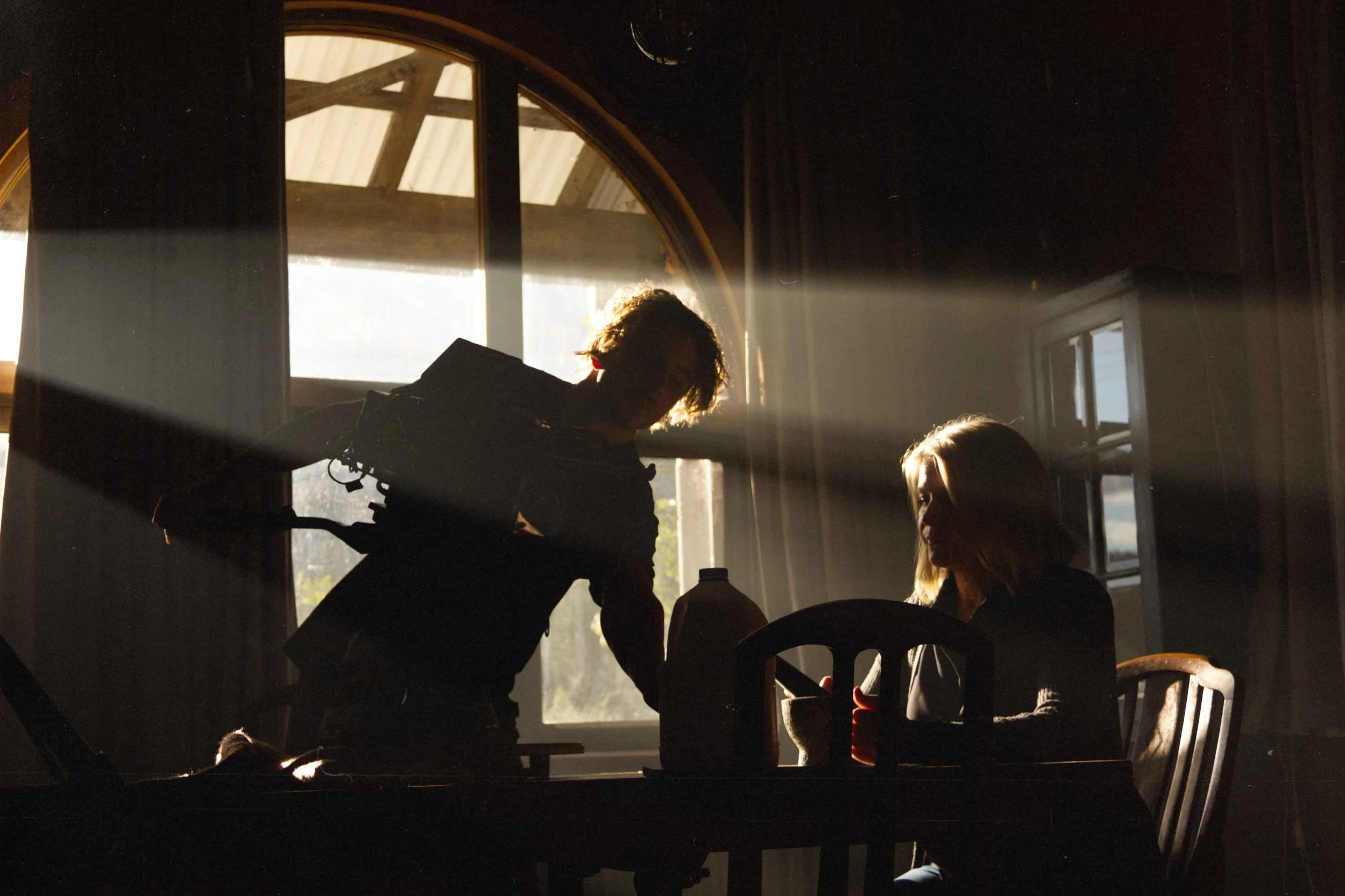 BTS of a short film called Lilium, directed by Musashi Wakaki. A dark interior scene with two women sitting at a dining table and a man standing, all silhouetted against sunlight streaming through a large arched window with wooden framing.