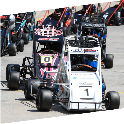 A lineup of small race cars at a race track, with cars numbered 0 and 1 prominently in the front, surrounded by other similar race cars.