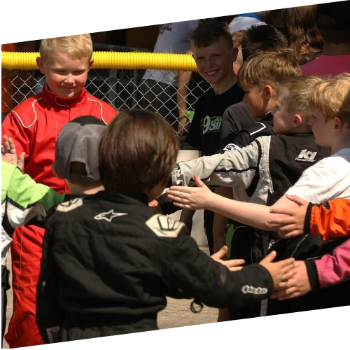 Children at a quarter midget race gathering near a fence, some in racing suits, engaging in conversation and interaction.