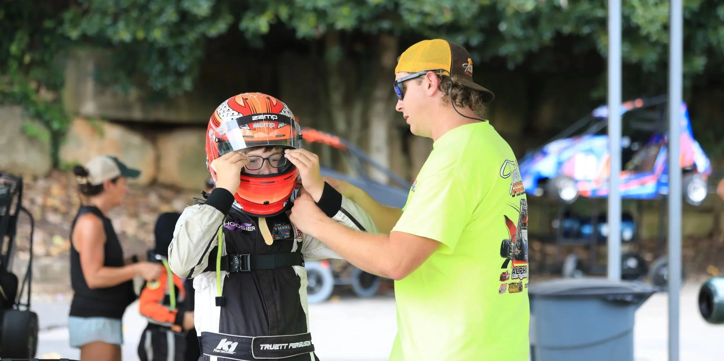 A race car driver in racing gear adjusts his helmet while a man in a yellow T-shirt assists him. Other people and race cars are visible in the background.