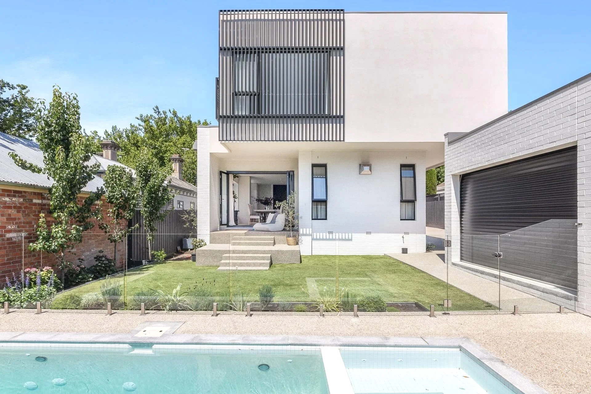 Modern two-story house with a backyard, features a glass fence, a pool in the foreground, a small lawn with plants, a patio with outdoor furniture, and architectural details including black slatted balcony panels, white walls, and a garage with black roller door.