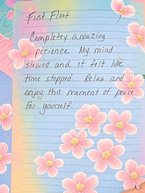 A handwritten note on lined paper surrounded by pink flowers on a blue background. The note reads: 'First Float Completely amazing experience. My mind slowed and it felt like time stopped. Relax and enjoy this moment of peace for yourself.'